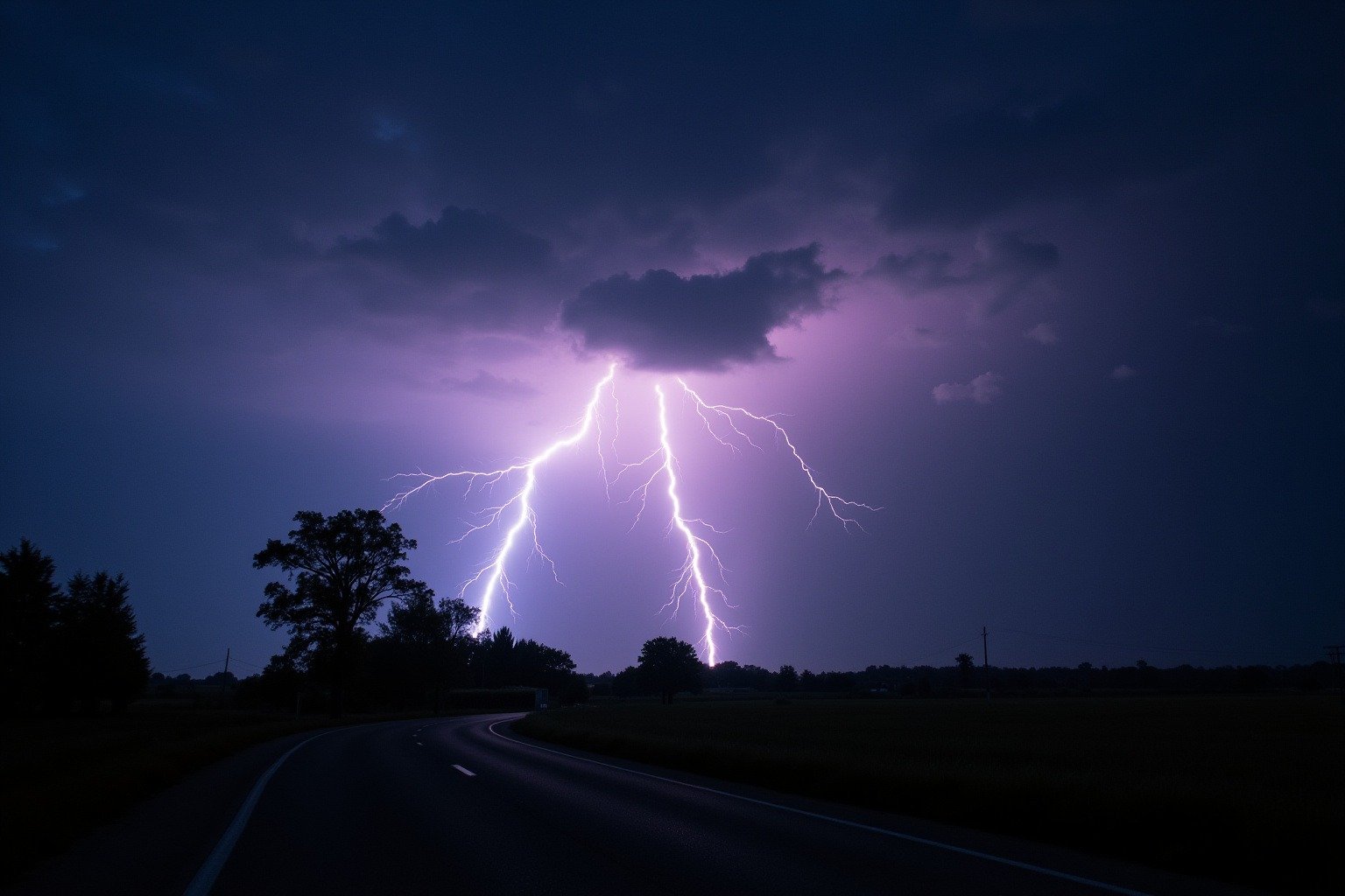 Distant heat lightning illuminating towering cumulonimbus storm clouds over a dark rural horizon at twilight with tree silhouettes in the foreground